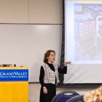 Female student presenting at the front of a carpeted conference room, pictures of a forest projected behind her